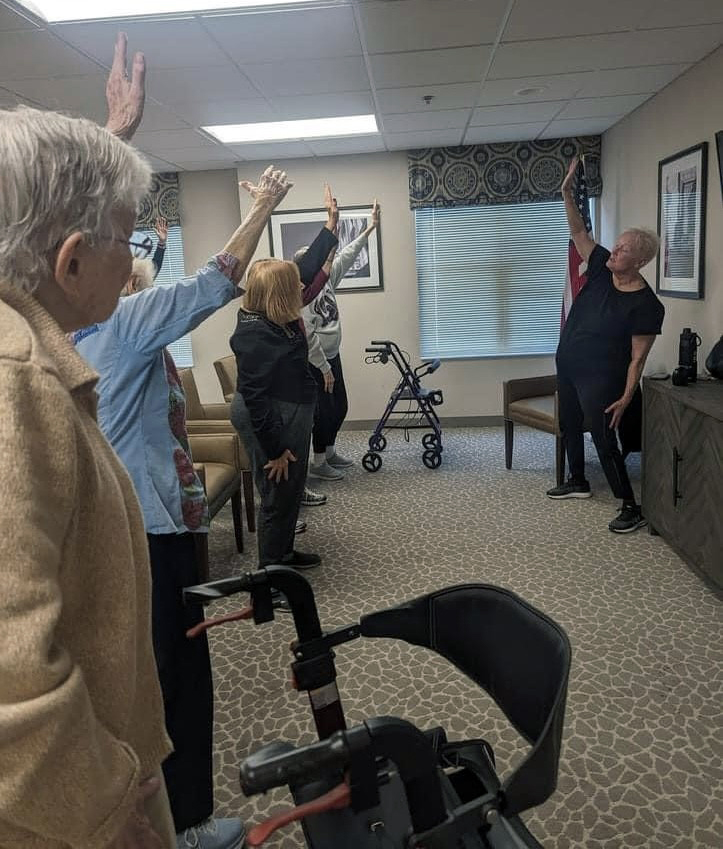 A group of elderly people in a room with their arms up, participating in an exercise session with a volunteer instructor in The Neighborhood.