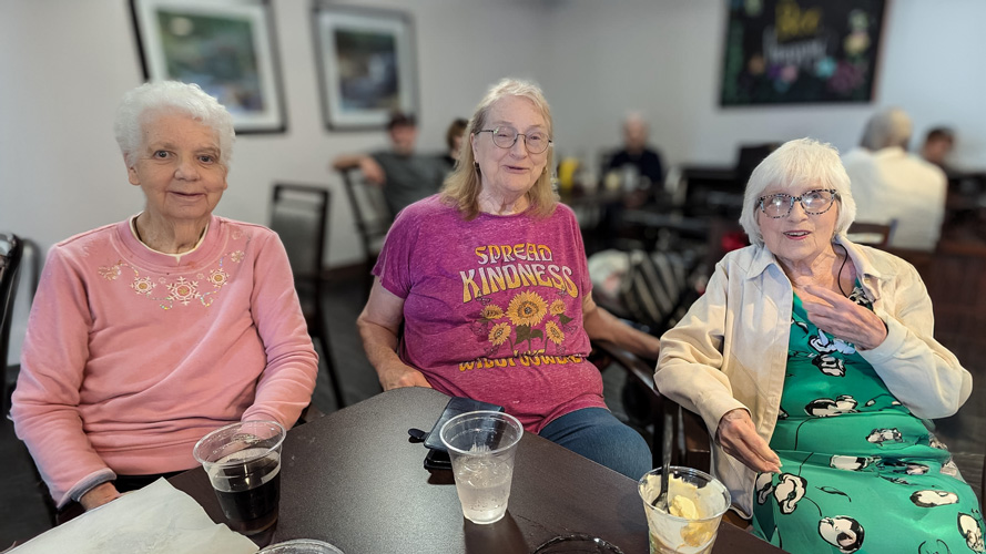 Three senior women sit together at a table in a social setting, enjoying drinks and ice cream, each with a cheerful expression, fostering a sense of community.