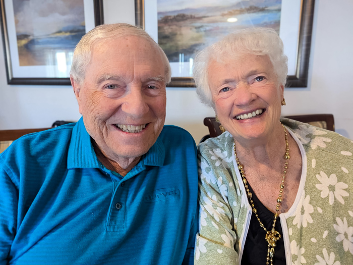 A senior couple with warm smiles sit closely together, creating a heartwarming moment in a room adorned with framed landscape paintings.