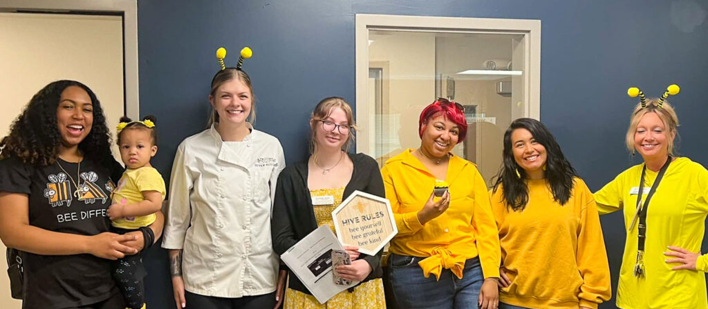 A group of six women, including one holding a toddler, dressed in bee-themed attire with yellow clothing and bee antennae headbands, smiling and posing together during a senior living community event.