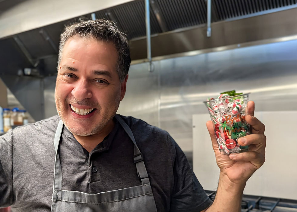 A smiling man in a gray shirt and apron holding a festive cup filled with holiday treats, celebrating Christmas in July in a senior living community kitchen.