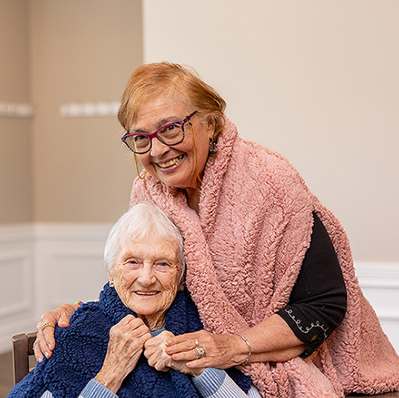 Two senior residents smile warmly while wrapped in cozy vests, one in blue and the other in pink, sharing a joyful moment together in a senior living community.
