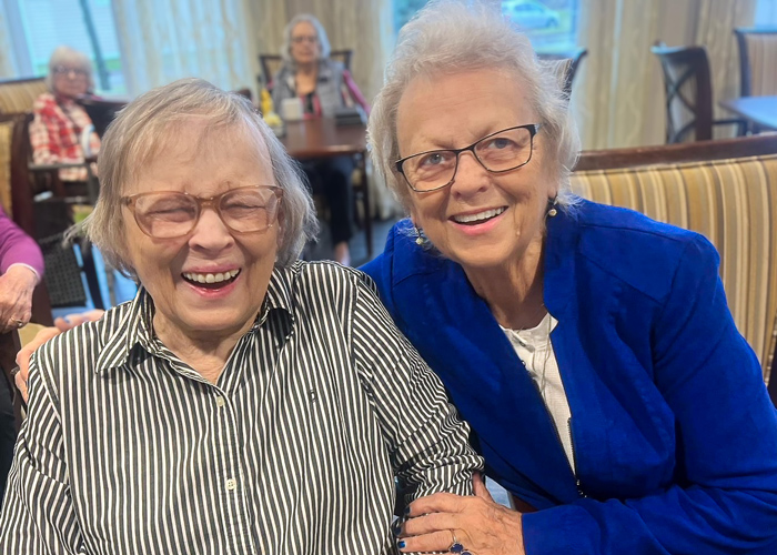 Two senior residents share a joyful moment together, smiling warmly in a dining area. One wears a striped shirt, and the other sports a bright blue blazer.