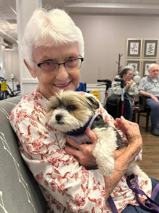 A senior resident with short white hair and glasses smiles while holding a small, fluffy dog in her arms at a senior living community, creating a heartwarming and comforting moment.