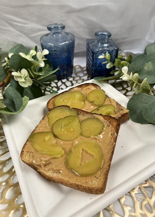 Toasted sandwich bread topped with creamy peanut butter and pickle slices, plated on a white dish with greenery and blue glass decor in the background.