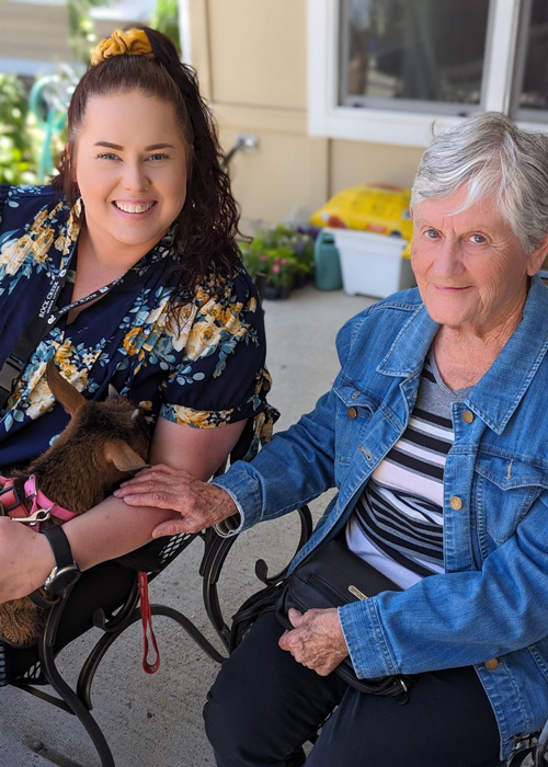 Libby and a senior resident sitting outside at Rock Creek Senior Living, smiling and gently petting a baby goat during a warm, sunny visit.