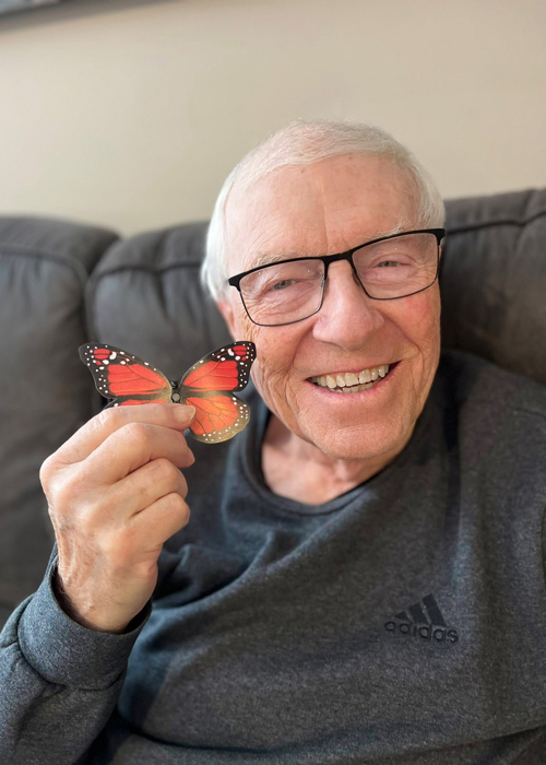 Senior resident smiles while holding a vibrant butterfly decoration, enjoying a relaxing and cheerful moment during a creative or seasonal community activity.