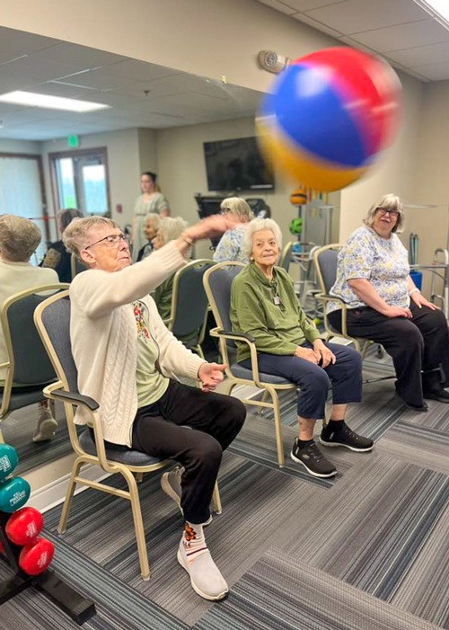 Senior residents play beachball volleyball while seated in chairs, staying active and social in a lighthearted wellness event with team members nearby.