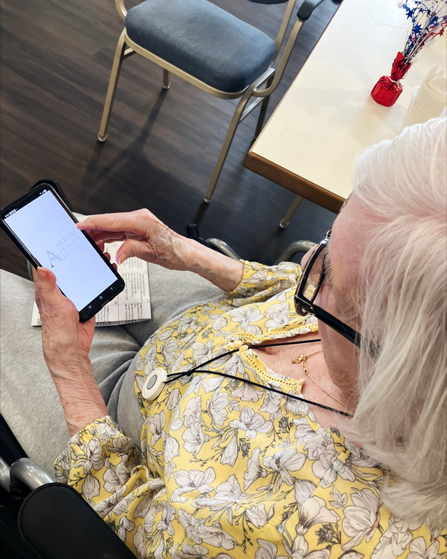 Senior resident in floral blouse sits at a table using a smartphone to open the Arrow Senior Living app, engaging with technology in a supportive environment.