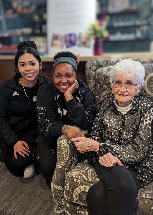 Two team members crouch beside a smiling senior resident seated in a patterned armchair, capturing a joyful moment.