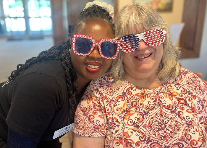 Two women smile brightly while wearing oversized patriotic sunglasses, enjoying a festive moment together at a community celebration.
