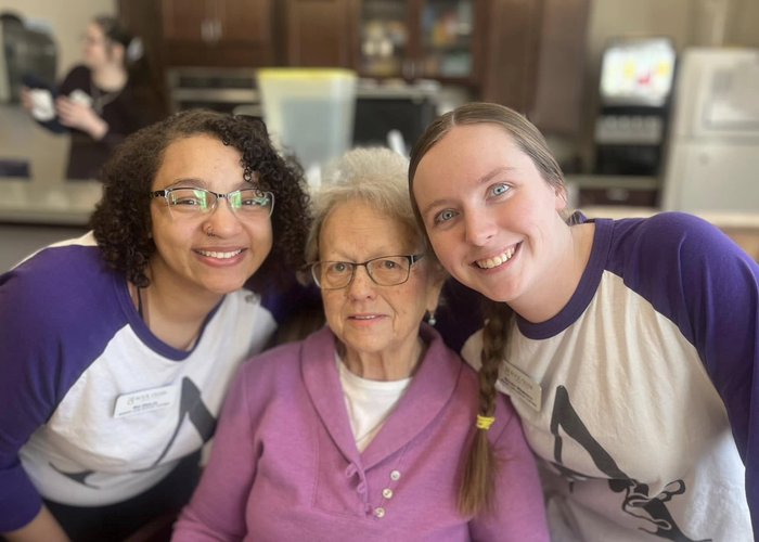 A senior woman in a lavender sweater smiles while seated between two cheerful care team members in matching purple shirts at a welcoming senior living community.