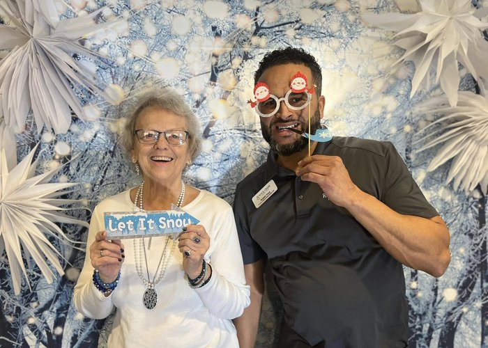 Senior woman and team member pose playfully in front of a winter-themed backdrop, holding holiday props including a “Let It Snow” sign and festive glasses.