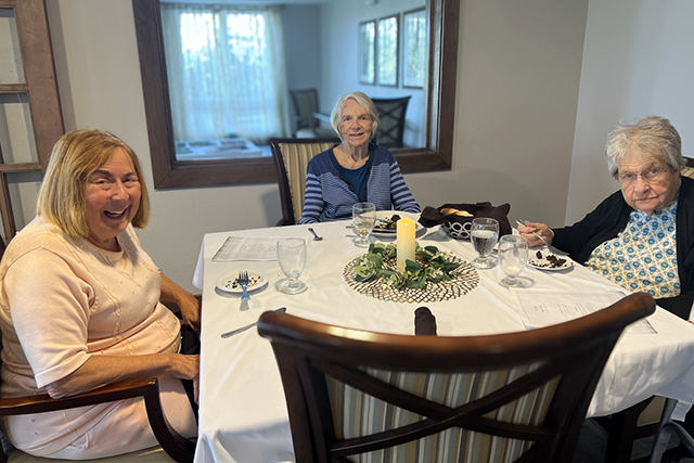 Three senior women sit together at a dining table, smiling and enjoying dessert in a warmly lit senior living dining room.