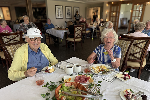 Senior couple at a dining table share a meal with coffee and dessert, surrounded by fellow residents.