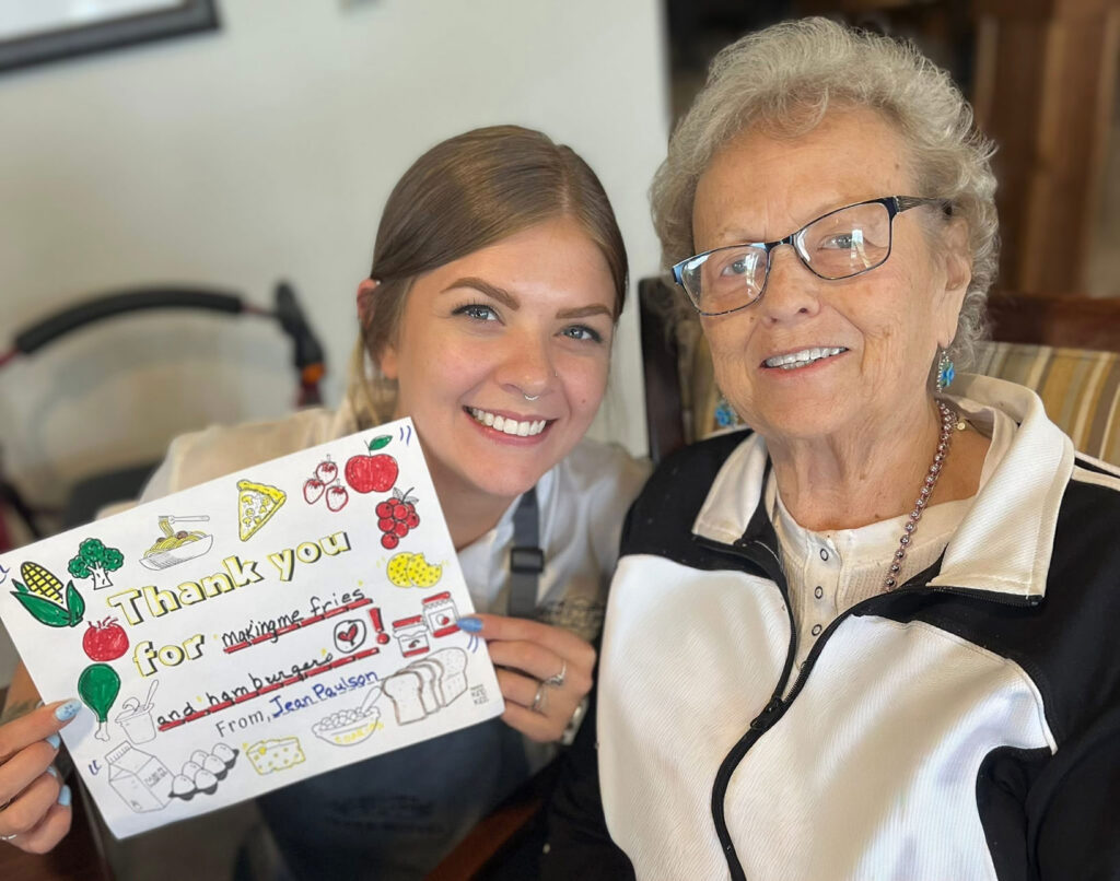 Senior resident and culinary director smile together while holding a colorful thank-you note decorated with drawings, showing gratitude for meals and care provided.