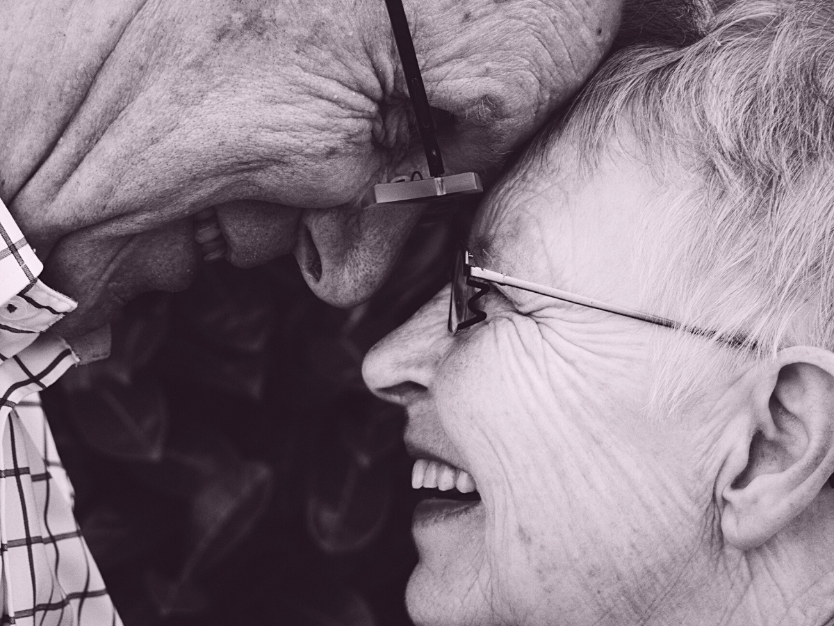 A joyful senior couple share an intimate moment, smiling as they press their foreheads together while wearing glasses, capturing love, connection, and happiness in close-up.