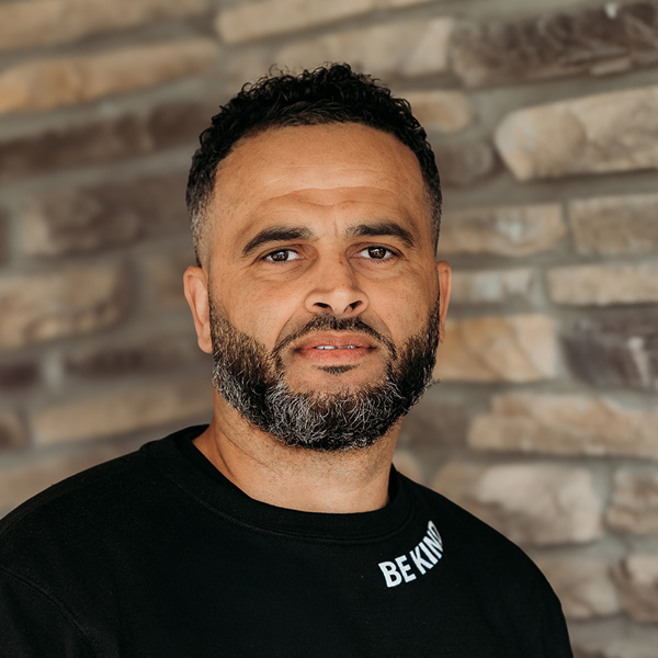 Robert Tighe, Plant Operations Director at Rock Creek Senior Living, smiling in a professional headshot. He has short curly hair, a trimmed beard, and wears a black shirt with the phrase “Be Kind,” standing in front of a stone wall background.