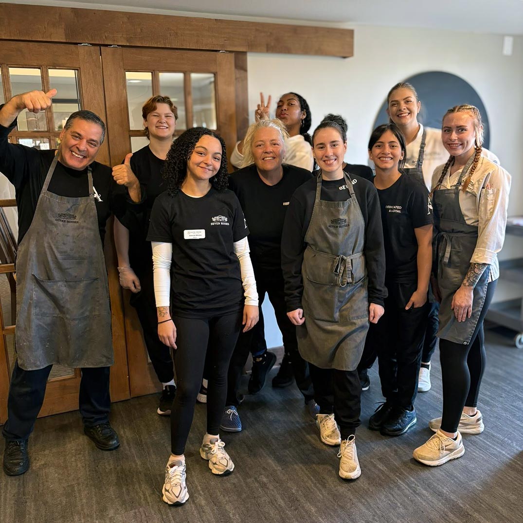 A group of culinary team members wearing black shirts and aprons smile and pose together in the community dining space.