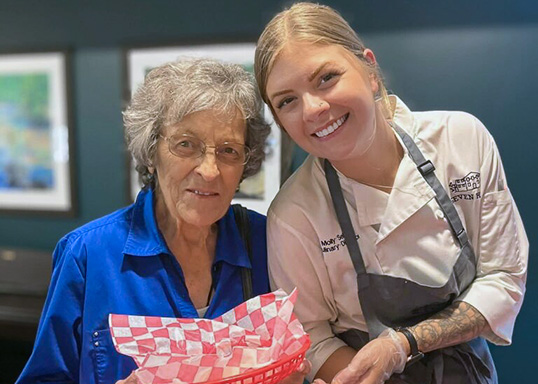 A senior woman smiles joyfully with Rock Creek's Culinary Director, holding a red fry basket during a community event.