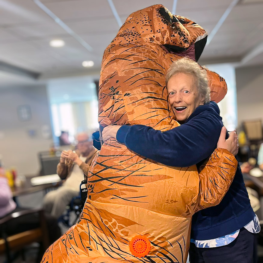 A senior woman and a team member in an inflatable t-rex costume embrace.