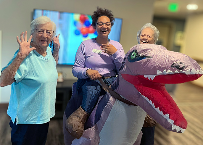 Senior residents smile alongside a team member wearing an inflatable dinosaur-riding costume.