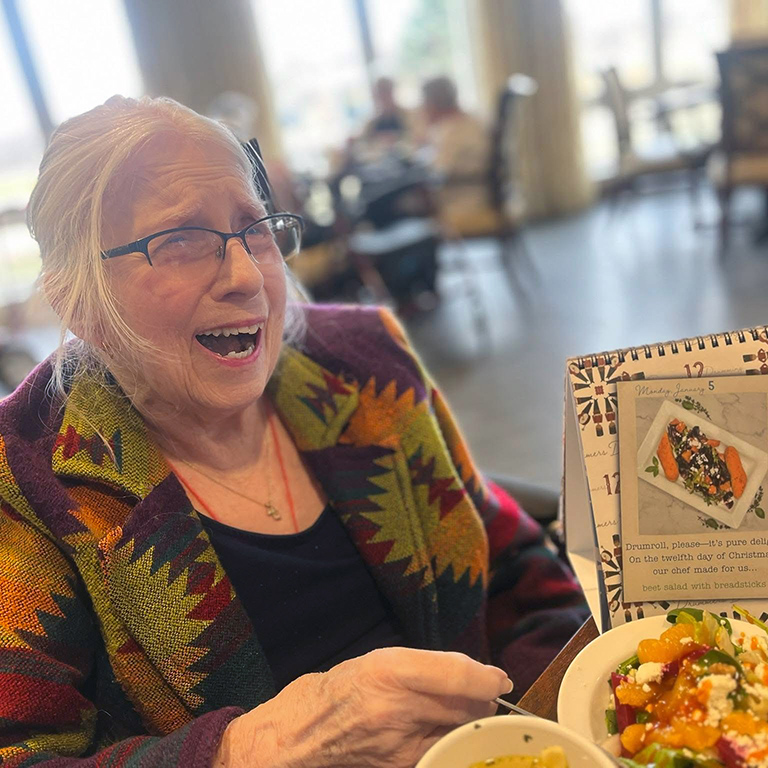 A senior woman smiles widely next to a themed chef-prepared meal.