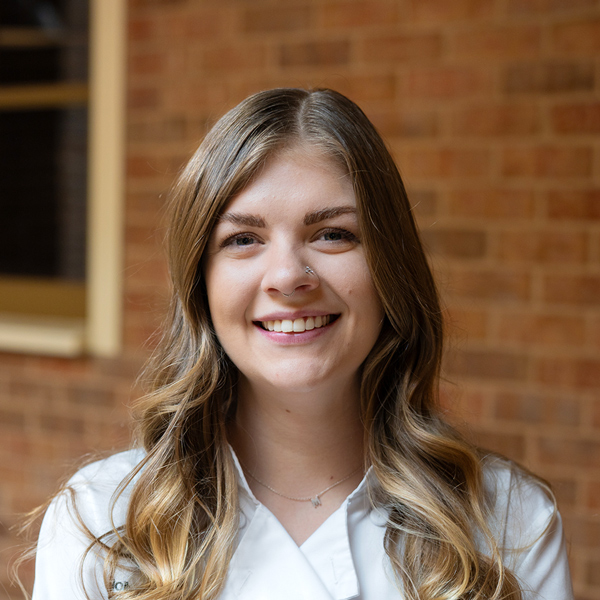 Molly Seher, Culinary Director at Rock Creek Senior Living, smiling in a professional headshot, wearing a white chef coat, standing in front of a softly blurred brick wall background.