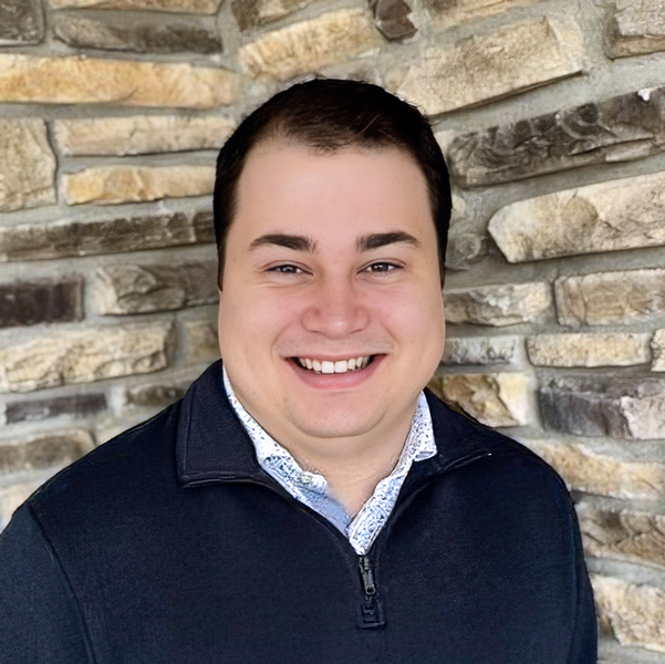Nick Nelson, Senior Living Director at Rock Creek Senior Living, smiling in front of a stone wall, wearing a dark quarter-zip sweater over a patterned collared shirt.