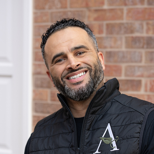 Robert Tighe, Plant Operations Director at Rock Creek Senior Living, smiling in a professional headshot, wearing a black vest, standing in front of a brick wall background.