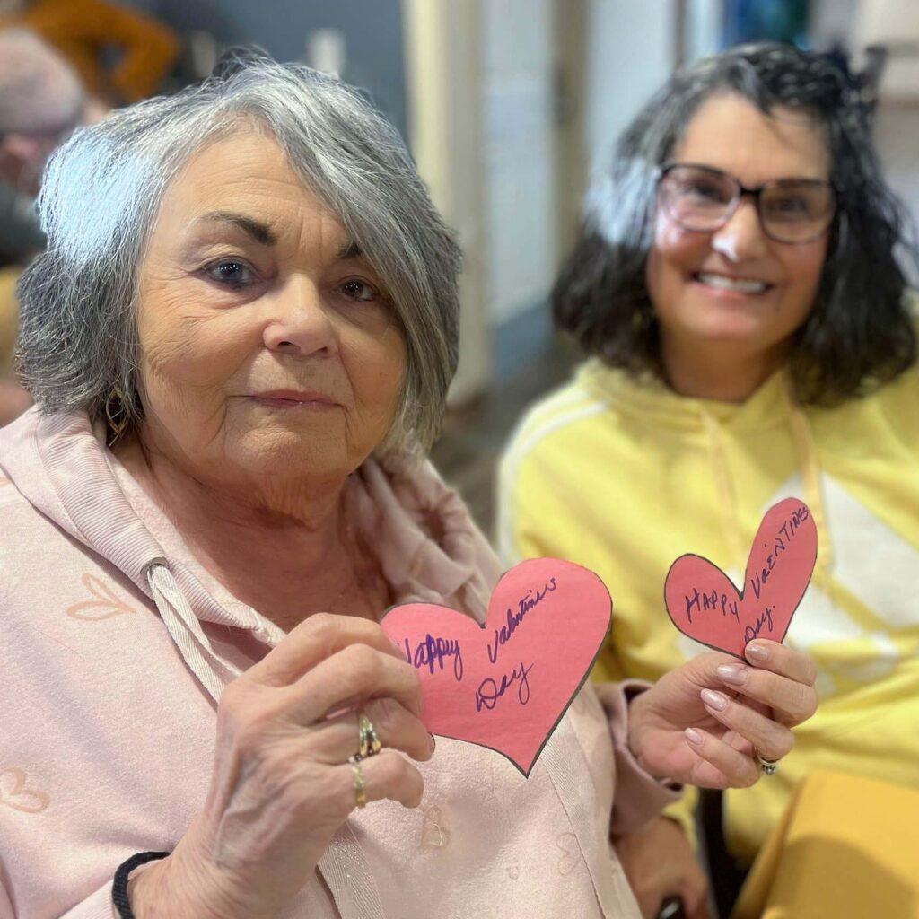 A loved one and senior resident smile, each holding a red paper heart.