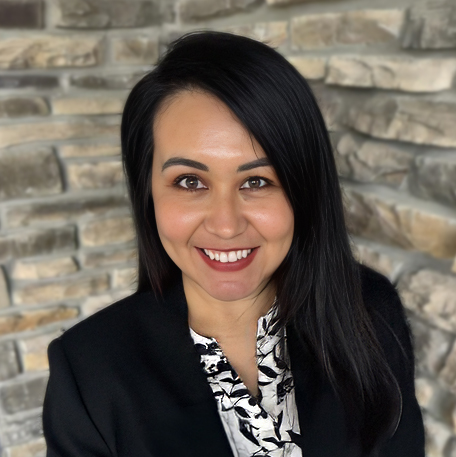 Esmeralda “Esme” Castil, Resident Services Director at Rock Creek Senior Living, smiling in a professional headshot with long dark hair, a black blazer, a patterned blouse, and a stacked stone wall background.