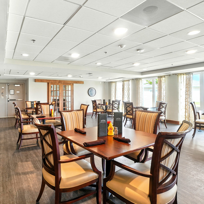 Spacious senior living dining room with wood tables, cushioned seating, and large windows filled with natural light.