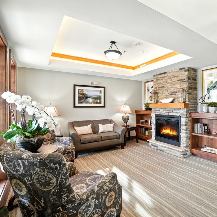 Cozy senior living lounge with fireplace, comfortable seating, bookshelves, and warm natural lighting.