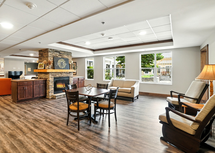 Bright senior living lounge with fireplace, seating areas, large windows, and wood flooring for resident relaxation.