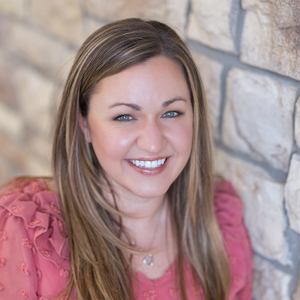 Stacey Russell, Regional Executive Director at Arrow Senior Living, smiling in a professional headshot. She has long light brown hair, wears a pink textured blouse, and is posed against a tan stone wall background.