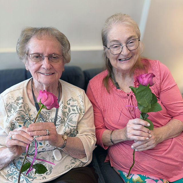 Two senior women smile brightly at Rock Creek Senior Living, seated together, each holding a long stem rose.